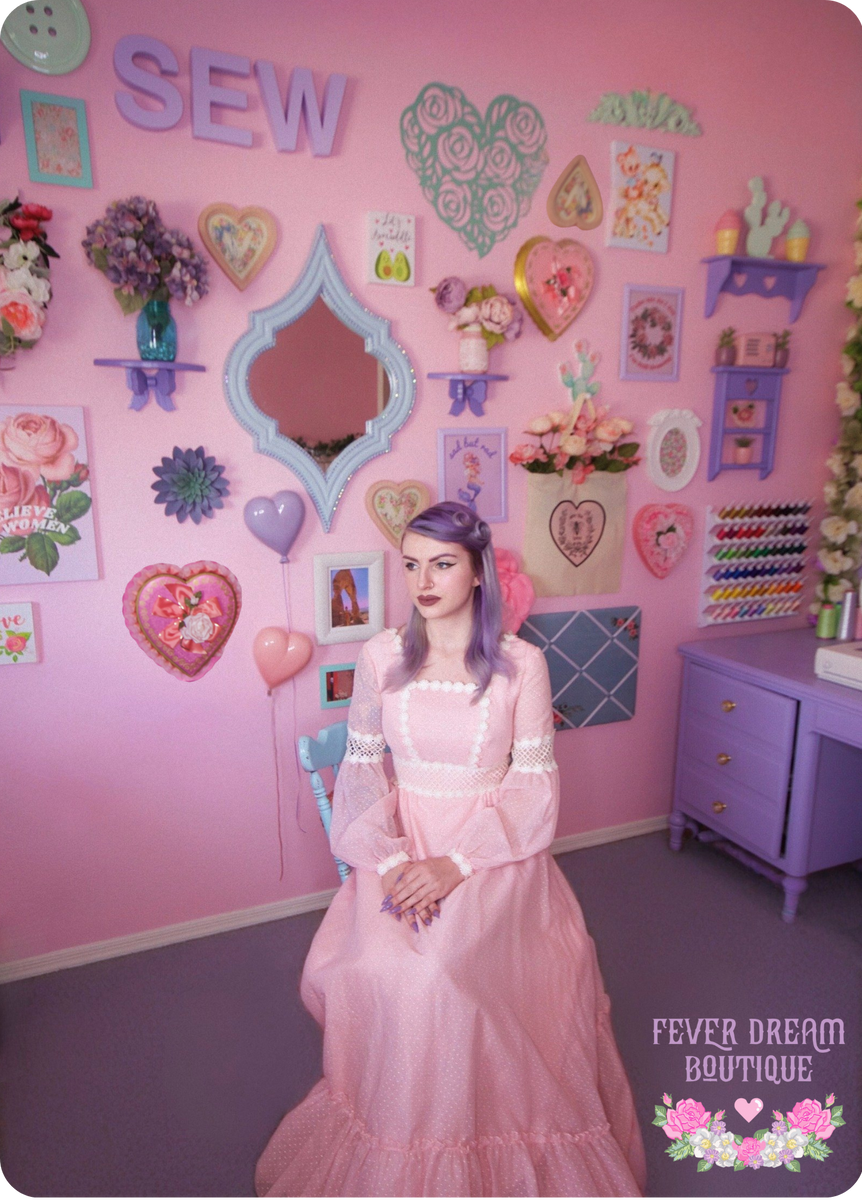 Emily Eileen, the designer and creator behind Fever Dream Boutique is sitting in a pink victorian dress. She is in a pink sewing room with lavender carpet. There is a gallery wall behind her with various vintage and thrifted trinkets.